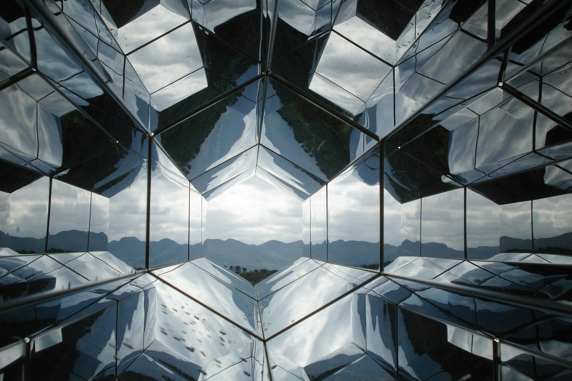 Kaleidoscopic structure of reflective glass panels seen from inside, with a cloudy sky and mountains at the centre